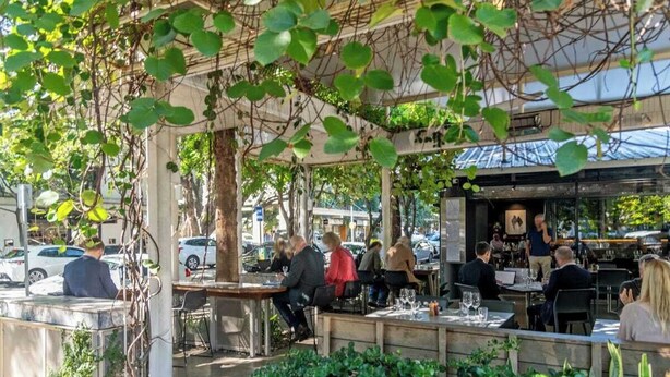 Un espacio al aire libre con mesas y sillas, rodeado de vegetación y una estructura de pérgola.