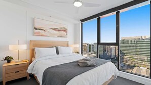 Una habitación de hotel moderna con una cama grande, una mesita de noche con lámpara, ventilador de techo y vistas a la ciudad a través de amplios ventanales.