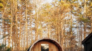 Eine Holzhütte mit rundem Design, einer Tür und einem Fenster, umgeben von einem Kiesweg und einem Wald.