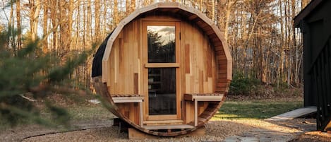 Una cabaña de madera con diseño circular, una puerta y un ventanal, rodeada por un sendero de piedra y bosque.