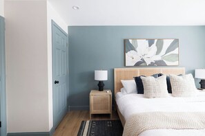 Une chambre à coucher comprenant un lit, des tables de chevet, une commode, une œuvre d’art accrochée au mur et une porte.