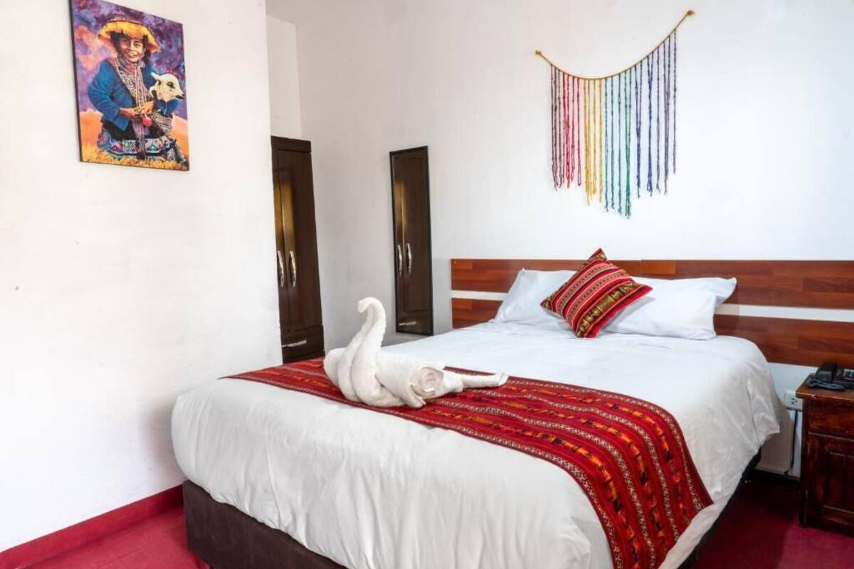 Habitación de hotel con cama, cabecero de madera, manta de diseño rojo y blanco, decoración de cisne blanco, tapiz de colores y un cuadro en la pared.