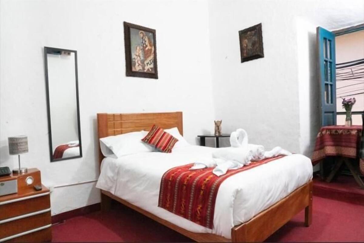 Una habitación de hotel con una cama de madera, ropa de cama blanca, un plaid rojo, un espejo y una pequeña mesa con una lámpara.