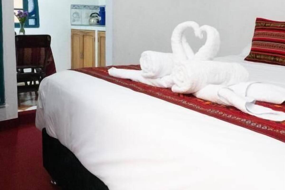 Una habitación de hotel con una cama bien hecha, una disposición de toallas en forma de corazón, un cojín a rayas rojo y blanco, y una mesita con un jarrón de flores.