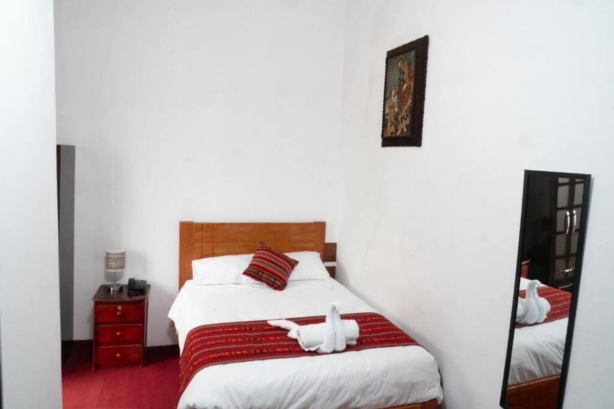 Una habitación de hotel con una cama de madera, ropa de cama blanca, una colcha roja con estampado, una mesita de noche con lámpara y un espejo en la pared.