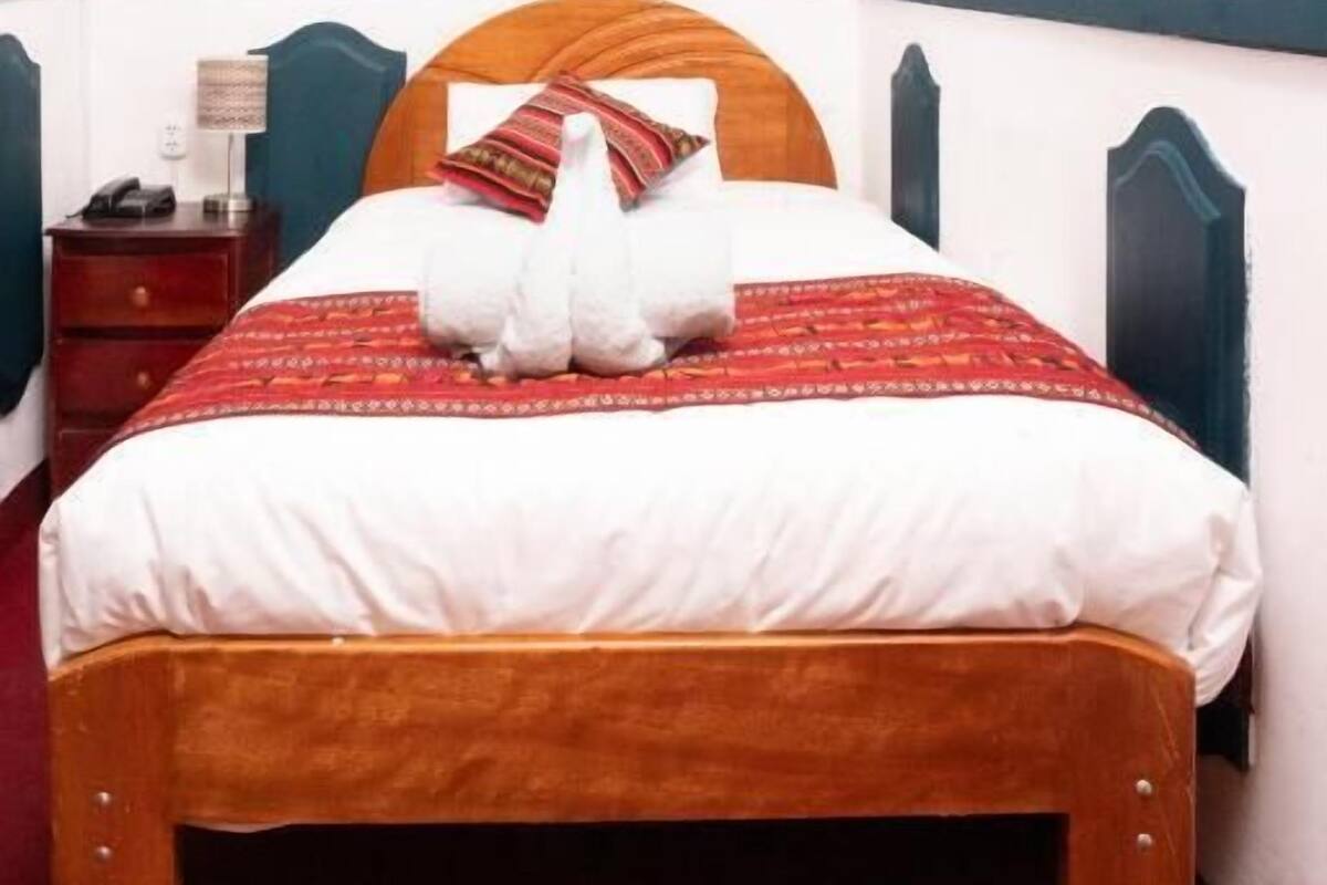 Una cama bien hecha con una colcha estampada en rojo y blanco, un cabecero de madera y una mesita de noche con una lámpara.