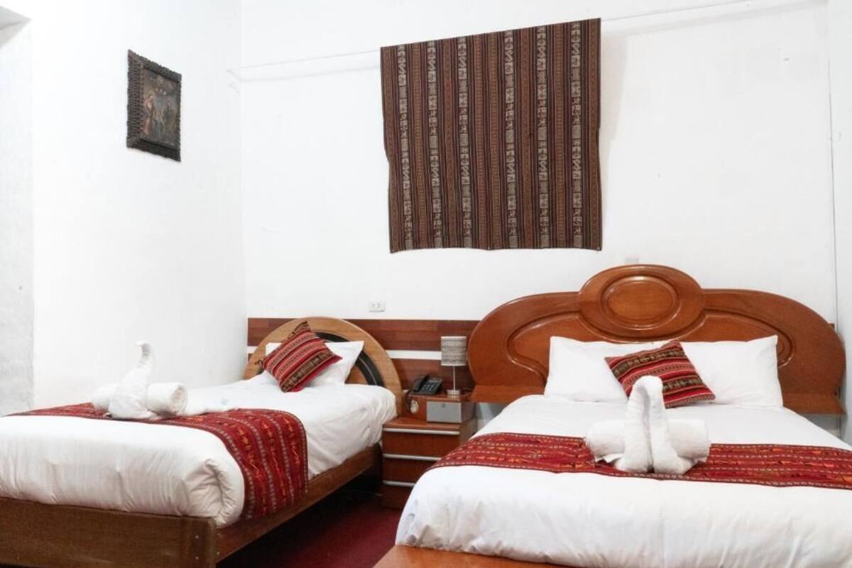 Una habitación de hotel con dos camas, cada una con cabecero de madera, ropa de cama blanca y mantas rojas estampadas. Hay un tapiz marrón a rayas sobre las camas.