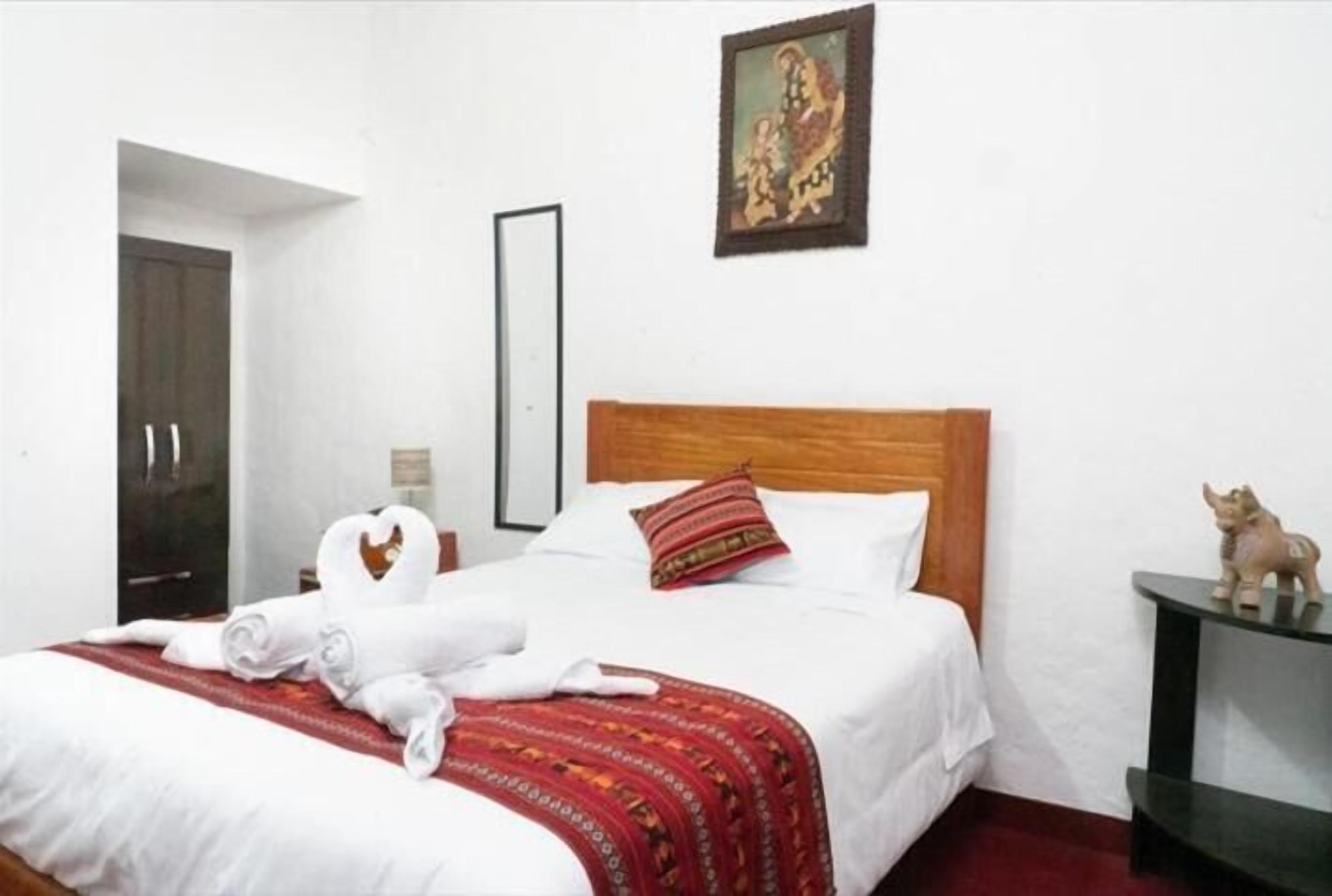 Habitación de hotel con una cama de madera, ropa de cama blanca, un plaid rojo con estampado y una pequeña estatua en una mesita de noche.