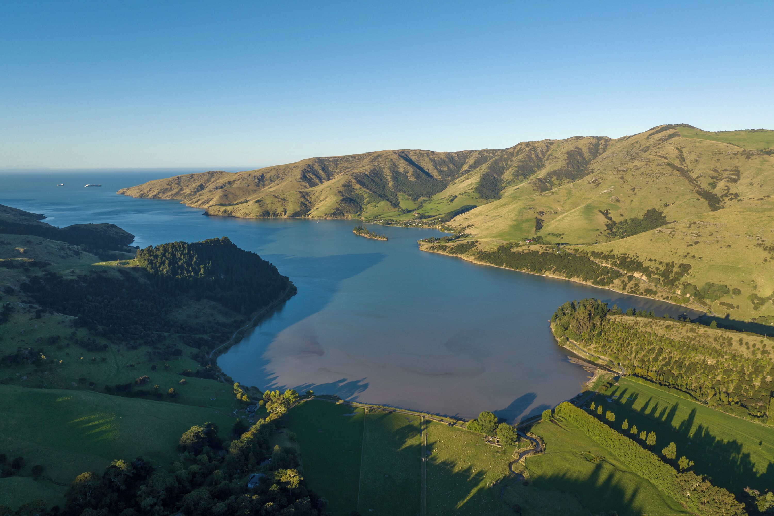 Vista aérea de un paisaje costero con una gran masa de agua, colinas verdes y un pequeño asentamiento.