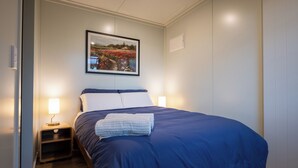 A neatly made bed with a blue cover, two bedside lamps, a framed picture on the wall, and a wooden nightstand. - Overleigh Eco-Pod (Port Levy)