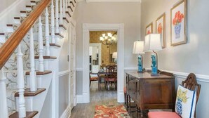 Un pasillo con escalera de madera, un comedor con candelabro y una alfombra decorativa.