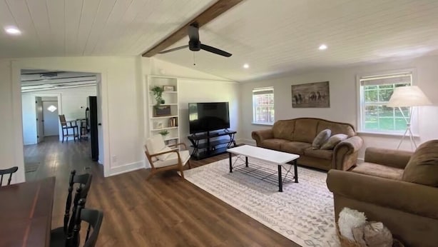 A spacious living room with a wooden floor, a ceiling fan, a TV, a sofa, and a dining area with a table and chairs.