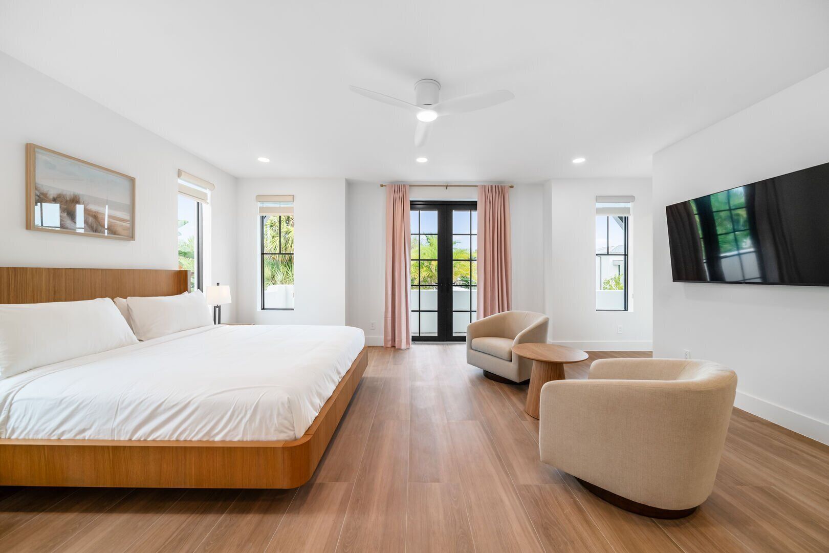 Quarto de hotel moderno com uma cama grande, área de estar com duas cadeiras, TV de tela plana na parede e grandes janelas com vista para o verde.