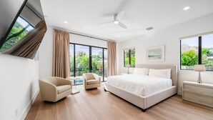 Une chambre moderne avec un grand lit, deux fauteuils, un ventilateur de plafond et de grandes fenêtres avec des rideaux.