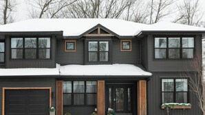 Une maison à deux étages, à l’extérieur sombre, avec un garage et un porche à l’avant doté d’une porte en bois.