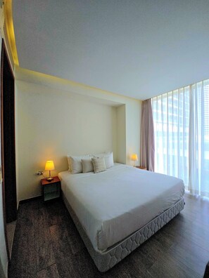 A hotel room with a large bed, two bedside tables with lamps, and a view of the city through sheer curtains. - A La Carte Da Nang 2-bedroom Sea View Apartment - Beach Front (Đà Nẵng)