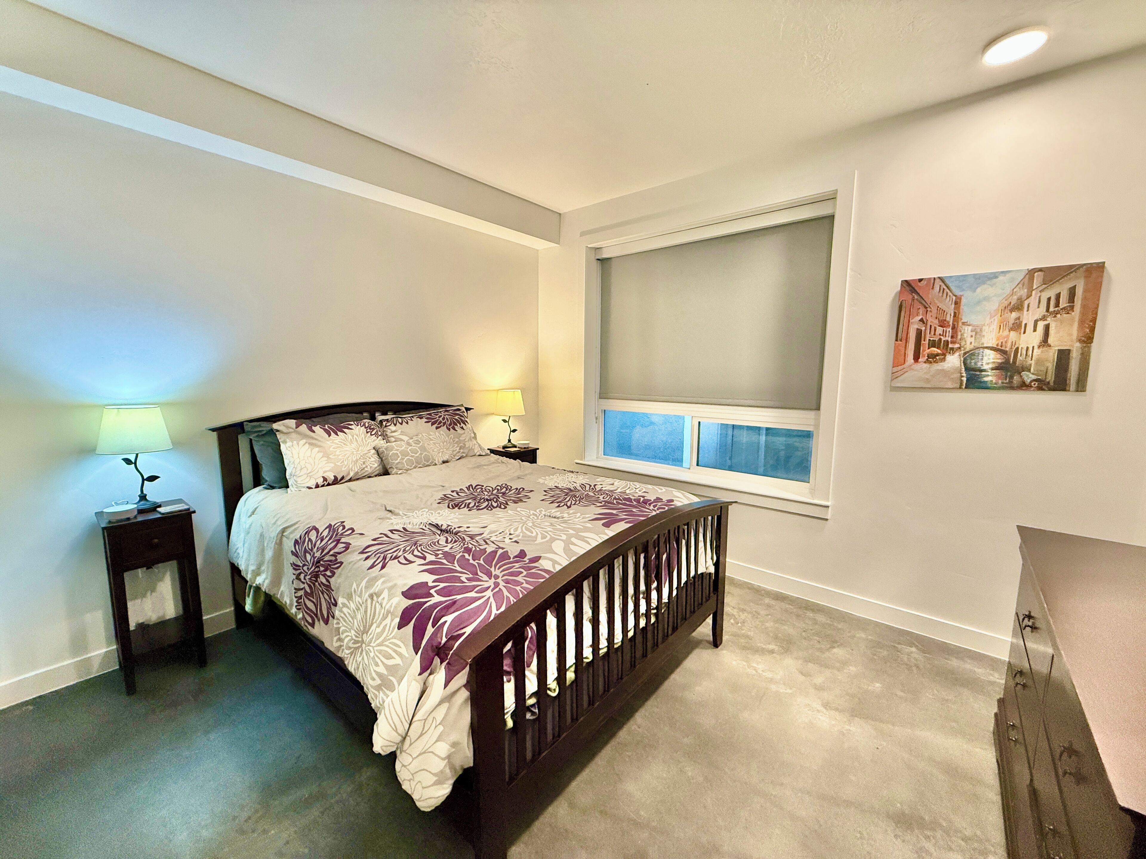 A bedroom with a bed, two bedside tables, a window with a view, and a painting on the wall.