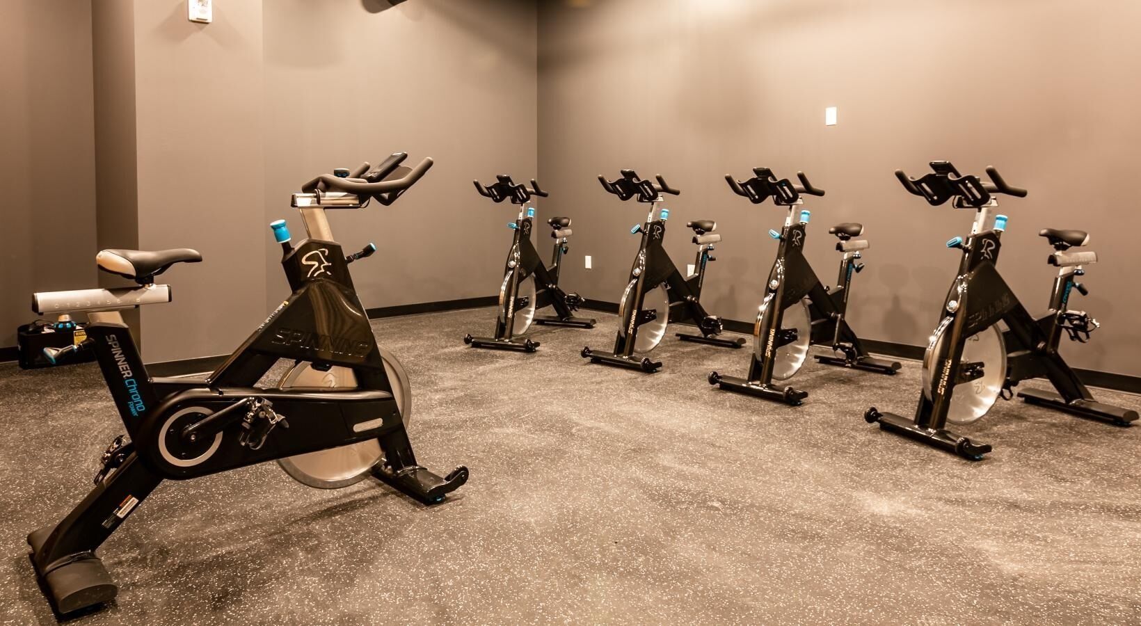 Várias bicicletas ergométricas em uma academia, com piso texturizado e paredes sem decoração.