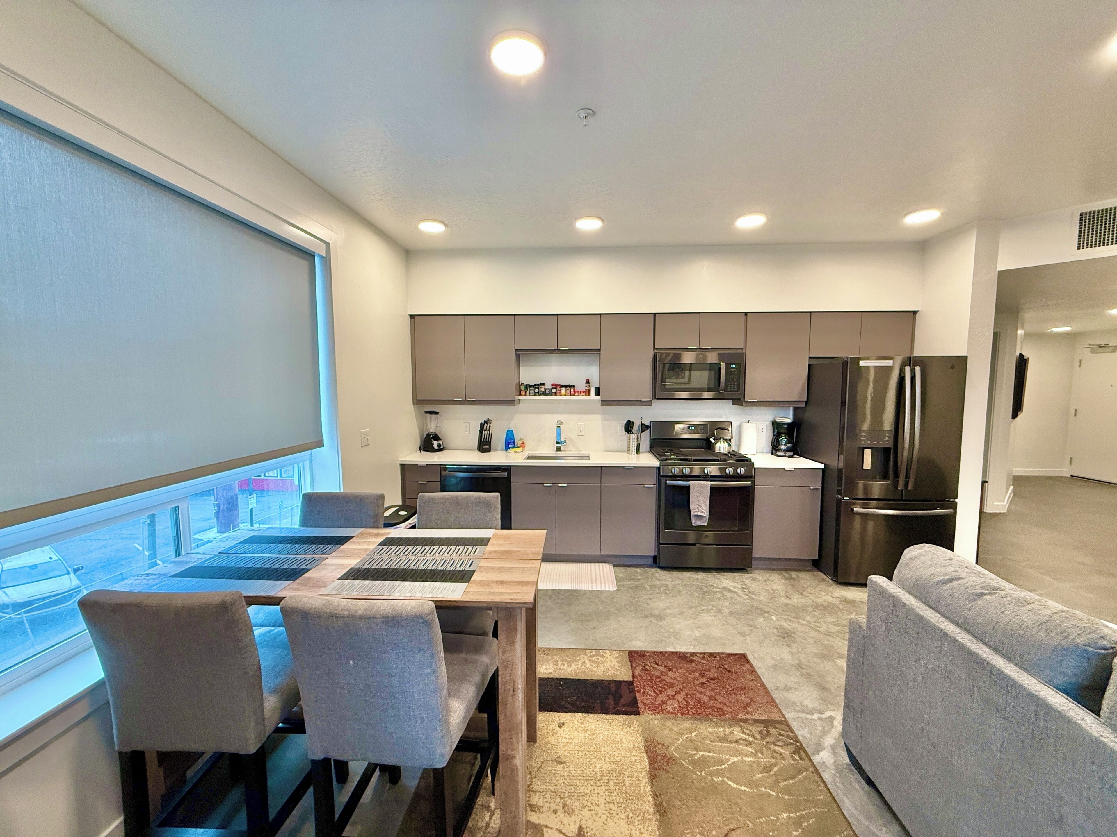 A modern kitchen with a dining area, including a table with chairs, a sofa, and a view through a window.