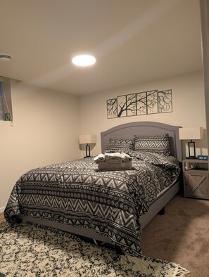 A neatly made bed with patterned bedding, a bedside lamp, and a clock on a wooden nightstand. - Modern Basement Suite w/ Free Parking & Trails (Winnipeg)