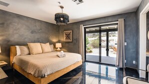 A modern bedroom with a large bed, a chandelier, and a view of the outdoors. - @ Marbella Lane - Zen-like Ambiance 3BR Home in JT (Joshua Tree)
