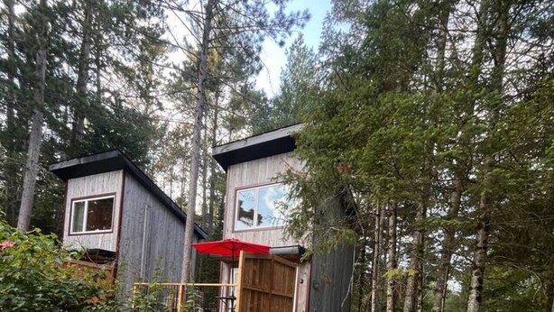 En hytte med to separate bygninger, hver med et vindue og en dør. Der er en terrasse med en rød parasol, og to Adirondack-stole står på en grussti.