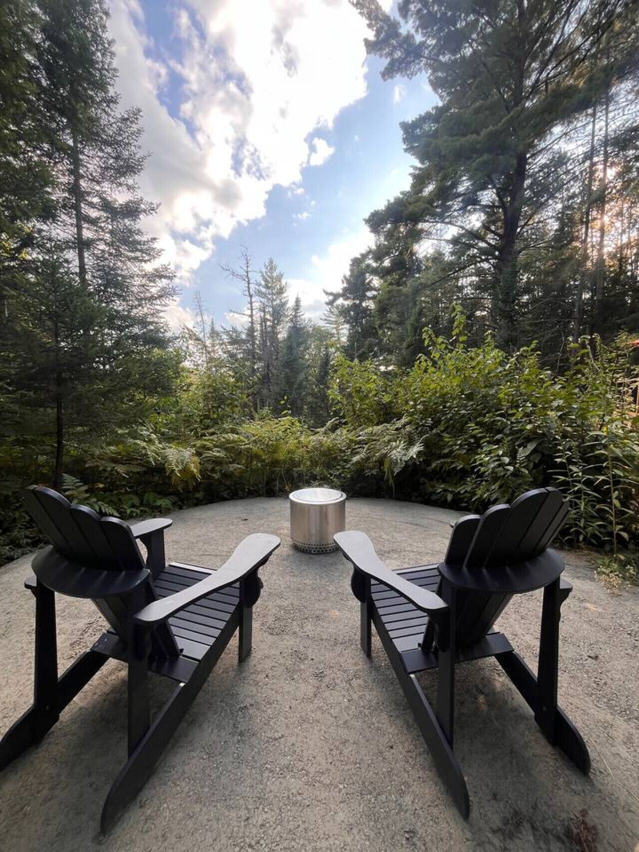 Two Adirondack chairs facing a circular fire pit in a forested outdoor area.
