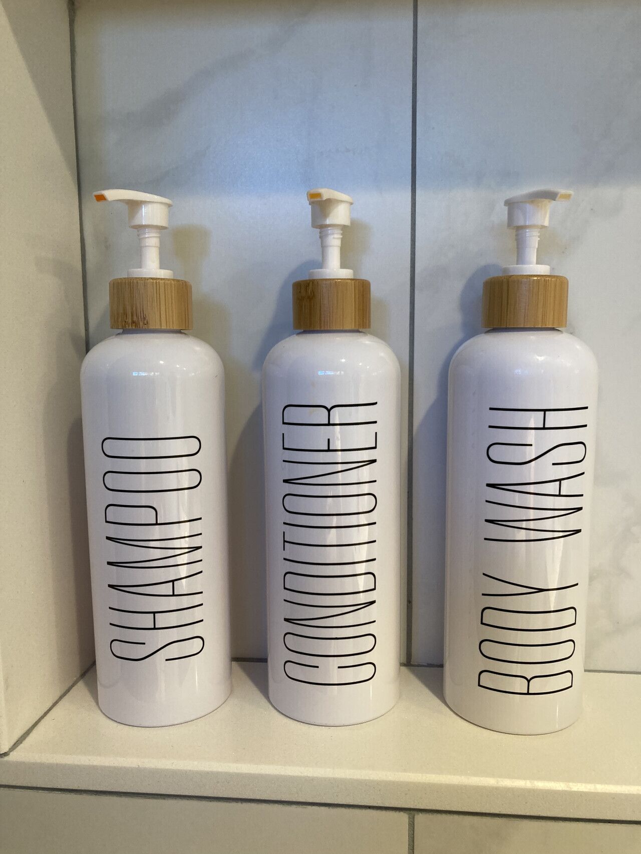 Trois bouteilles blanches, équipées de pompes et portant des étiquettes, sont posées sur une étagère.