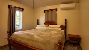 Une chambre avec un lit en bois, une fenêtre avec des rideaux, un appareil de climatisation et un petit banc en bois.