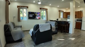 A modern living room with a black sofa and a recliner, a flat-screen TV mounted on the wall, and a kitchen area with wooden cabinets and a microwave. - Hidden Treasure Vacation Home (Caye Caulker)
