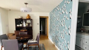 Ein Essbereich mit Holztisch, Stühlen und Teppich. An der Wand ist ein Vogelmuster zu sehen, und eine Tür führt in einen anderen Raum.