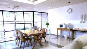 A modern dining area with a wooden table and chairs, a sofa, a kitchen area with appliances, and large windows offering a view of greenery. - HOTEL Reise Basis (Senboku)