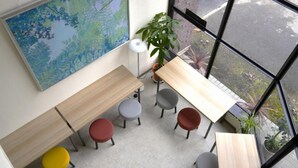 A modern office space with multiple wooden tables and colorful chairs, large windows offering a view of the street, and a large abstract painting on the wall. - HOTEL Reise Basis (Senboku)