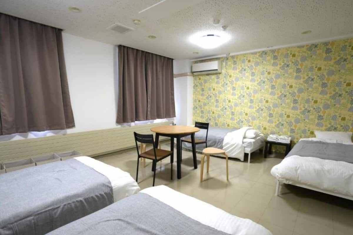 Habitación de hotel con dos camas individuales, una mesita con sillas, un escritorio y una silla. Se observa un equipo de aire acondicionado en la pared y una ventana con cortinas.