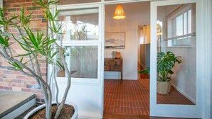Una habitación con pared de ladrillo, una planta en maceta, un banco de madera y una puerta que da a otra estancia.