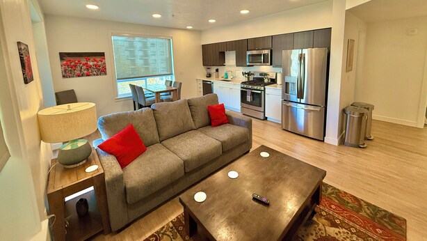 A modern living room with a grey sofa, a coffee table, and a dining area with a table and chairs. A kitchen with a refrigerator, stove, and microwave is visible in the background. - Luxurious New Condo in Downtown SLC (Salt Lake City)