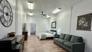 A spacious room with a bed, a green sofa, a wooden floor, a ceiling fan, and a wall clock.