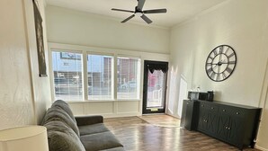 A spacious living area with a dark-colored sofa, a small table with a lamp, a ceiling fan, and a clock on the wall.