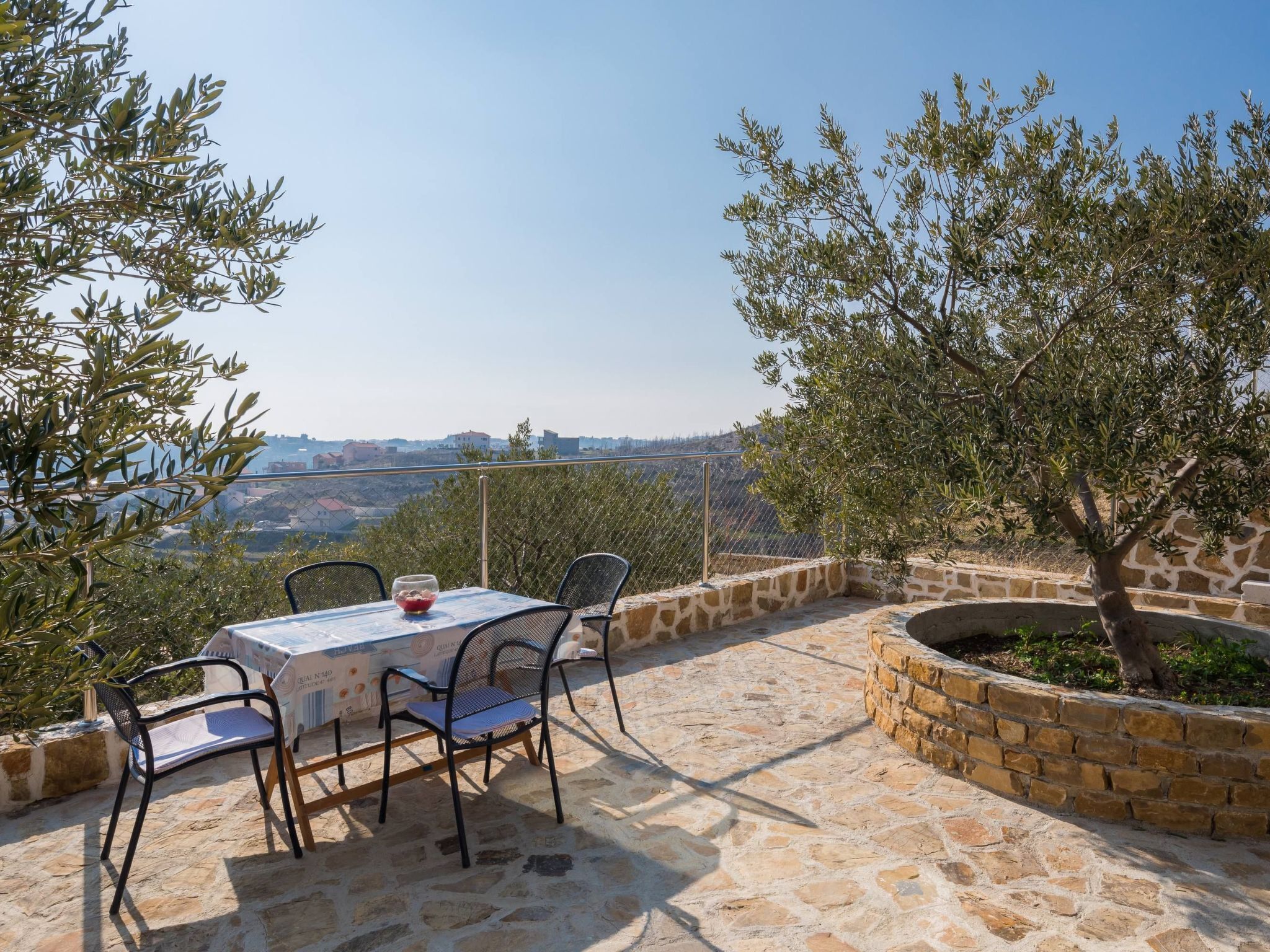 A stone patio with a table and chairs, olive trees, and a view of a city in the background.