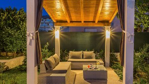 An outdoor seating area with a wooden pergola, cushioned seating, and a glass table with drinks. - in Stobreč mit privatem Pool by Interhome (Split/Stobreč)