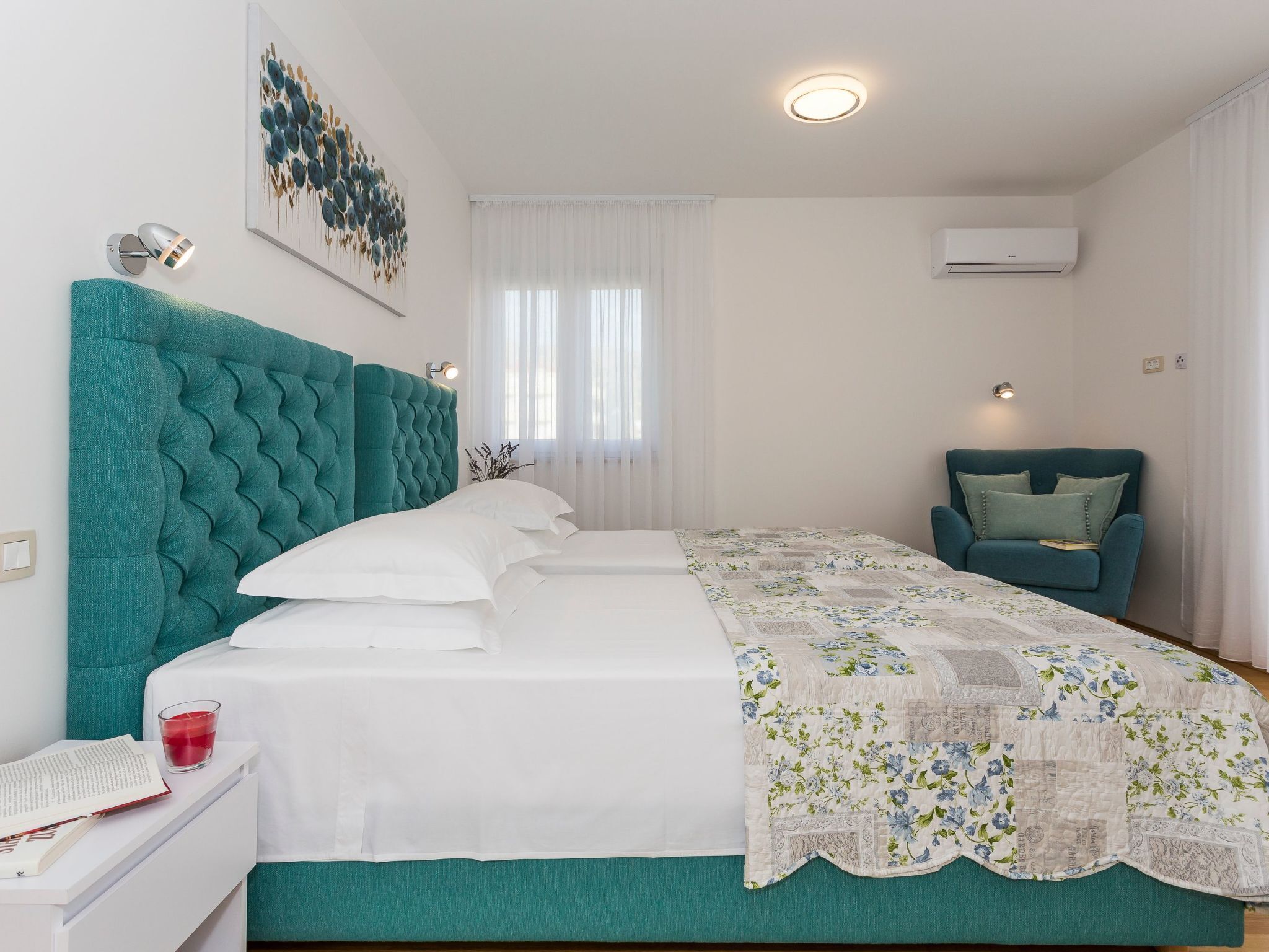 Camera da letto con una testiera imbottita color petrolio, un copripiumino bianco, una coperta a fiori, una poltrona color petrolio e un condizionatore d'aria a parete.