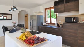 Eine moderne Küche mit Kochinsel, dunklen Holzschränken, einem Edelstahl-Kühlschrank und einem großen Fenster, das für natürliches Licht sorgt.