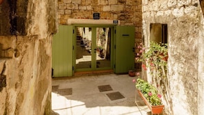 A stone building with a green door and shutters, a visible room with a bed and a window, and a small outdoor area with potted plants.