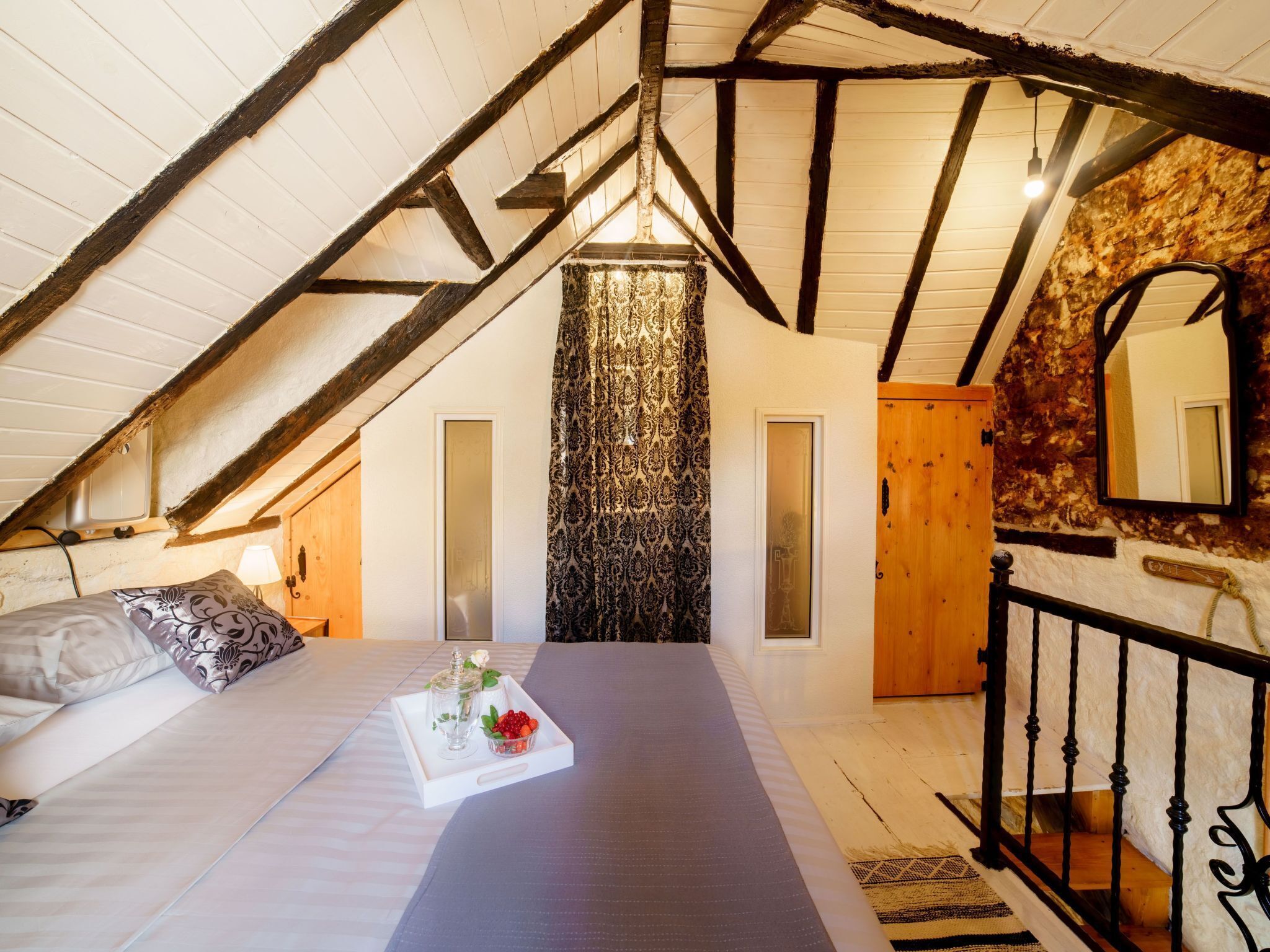 A bedroom with a sloped ceiling, a bed with a tray of fruit, a mirror, and a wooden door.
