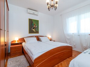 Quarto com cama de madeira, abajures, um quadro na parede, ar condicionado e uma janela com cortinas leves.