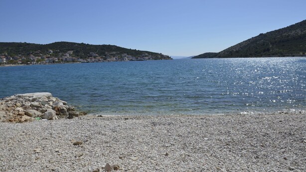 Una playa pedregosa con agua cristalina y un pueblo lejano en las colinas.
