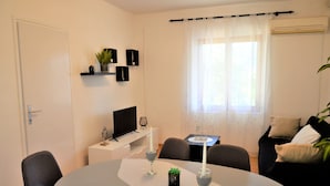 A modern living room with a dining table, chairs, a TV on a stand, a window with sheer curtains, and a sofa with cushions. - Erdgeschosswohnung, erste Reihe vom Meer by Interhome (Marina)