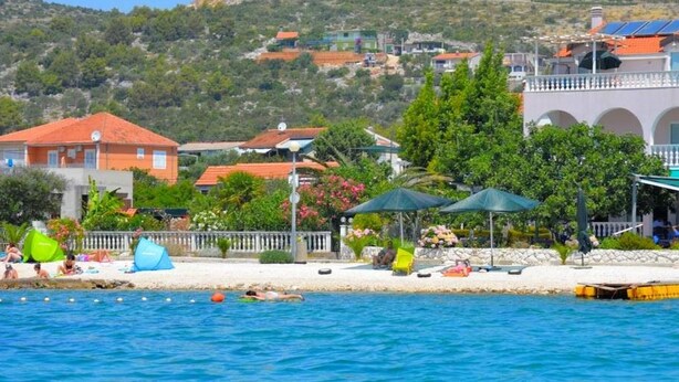 Un hotel costero con zona de piscina, sombrillas de playa y vistas al mar y edificios en la colina.