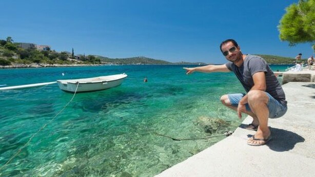 Ein Mann deutet auf ein Boot, das in klarem, blauem Wasser vor Anker liegt, mit einem Strand und einer fernen Küste im Hintergrund.