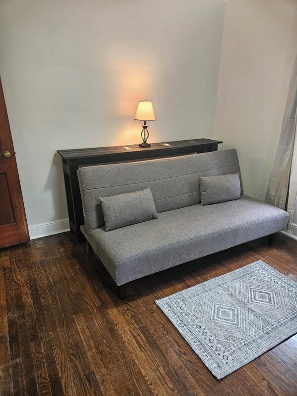 A grey sofa with two cushions, a small table with a lamp, a patterned rug, and wooden flooring.